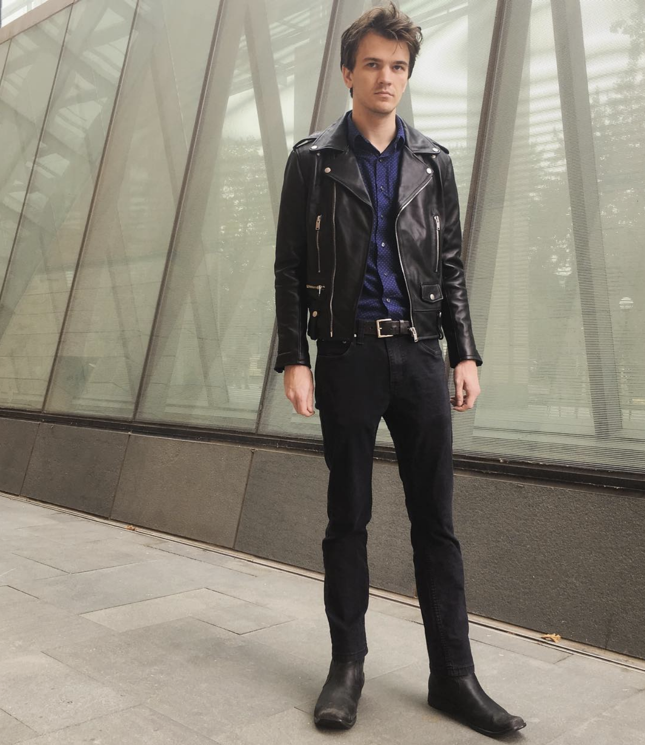 A photo of a man with brown hair wearing a black leather jacket and jeans standing in front of a glass structure.